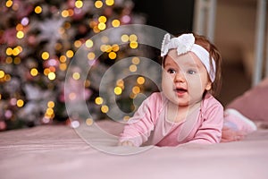 Infant baby girl with pink head band lying on belly and dreaming. Blurred decorated Christmas tree with lights in background.