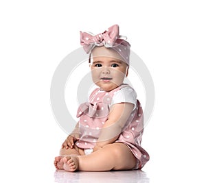 Infant baby girl toddler in polka-dot dress and headband with bow sits on the white floor looks at camera with interest