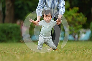 infant baby learn walking first step on grass field with mother holding hand helping in park