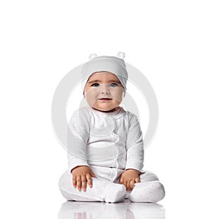 Infant baby toddler sits on the floor in white jumpsuit overall and funny hat with ears touching her legs