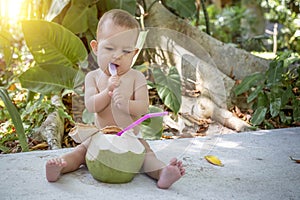 Infant baby at tropical vacation. Eats and drinks green young coconut. Sits on a ground and hold a spoon. Jungles on background