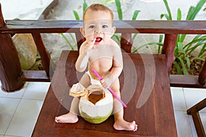 Infant baby at tropical vacation. Eats and drinks green young coconut. Sits on a wooden table and gnaw spoon. Teething