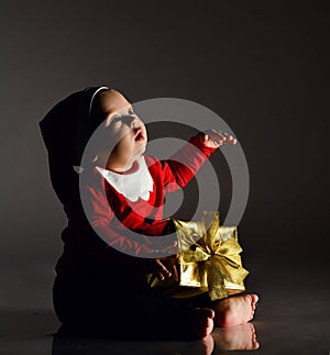 Infant boy toddler in santa claus costume is sitting with new year gift box present holding hand up, slapping, looking