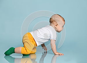 Infant child baby boy kid in yellow pants and white t-shirt is crawling away on all fours on blue background