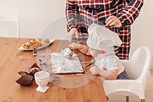 Infant cook baby portrait wearing chef hat playing with dough at kitchen table.