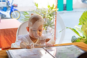 Infant girl is sitting on a baby`s high chair in a street cafe. Children reading chooses and studies the menu in the restaurant