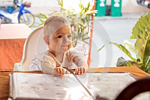 Infant girl is sitting on a baby`s high chair in a street cafe. Children reading chooses and studies the menu in the restaurant