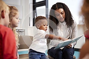 Infant school boy pointing in a book held by the female teacher, sitting with kids on chairs in the classroom, close up