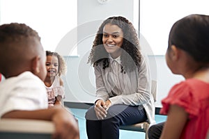 Infant school children sitting on chairs in a circle in a classroom talking to their smiling female teacher, close up, selective f