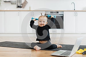 Infant in sportswear holding dumbbells near laptop on floor