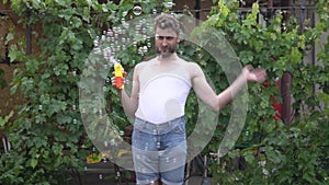 An infantile young man with a beard stands and launches soap bubbles from a toy bubble and waves it away with his hand