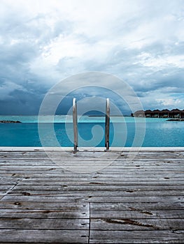 Infinite swimming pool ladder with wooden deck floor under blue cloudy sky on a beach of Indian ocean