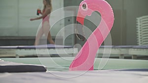 Inflatable rubber pink flamingo floating in a pool on the background of a passing beautiful girl in a bathing suit with