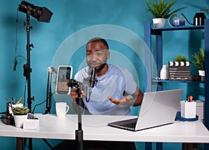 Influencer sitting down at desk with laptop in vlogging studio smiling in front of recording smartphone