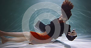 INFORMATION OVERLOAD. Cinematic side view, young business woman sinks under water with smart phone, laptop slow motion.