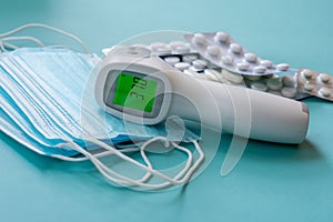 Infrared thermometer with digital green colored screen showing high body temperature on pile of medical masks and pills in blister