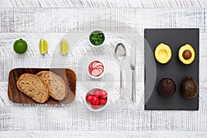 Ingredients for avocado toast. Slices  of fresh rye bread, lime, herbs, radish, avocados, spoon and knife on the white wooden tabl