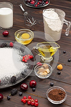 Ingredients for baking berry pie. Flour in black plate, cocoa powder. Measuring cup with flour, glass of milk, broken egg and salt