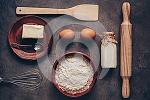 Ingredients for baking on a dark rustic background, flour, butter, eggs, rolling pin, whisk and paddle. Top view, flat lay