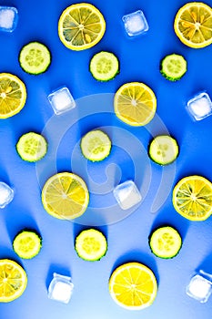 Ingredients for cooking cold spring lemonade drink with cucumber, pieces of lemon, ice cubes on blue background, top view