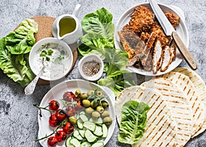 Ingredients for cooking greek gyros - chicken schnitzel, lettuce, tortillas, greek yogurt sauce, tomatoes, olives on a gray