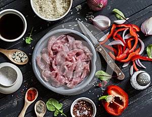 Ingredients for cooking meat stir fry with vegetables and rice - raw meat, sweet red pepper, red onion, rice, spices, on dark wood