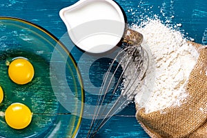 Ingredients for making the dough. A bowl of egg yolks, a hand whisk, flour and milk on the table. View from above