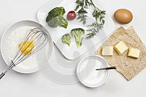 Ingredients for making dough. Broken egg with flour in bowl. Whisk on bowl. Butter on paper. Broccoli, tomatoes and dill in plate