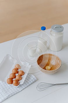 Ingredients for making dough or pasrty. Fresh eggs, milk and flour with whisk on white wooden kitchen table. Products for cooking