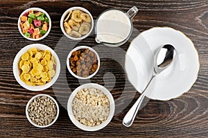 Ingredients for muesli dried fruits, corn flakes, oatmeal, sunflower seeds, banana chips, yogurt and bowl, spoon on table. Top