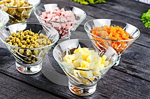 Ingredients shredded for salad in transparent plates on a black wooden background. Olives, pickled cucumbers, sausages, carrots, p