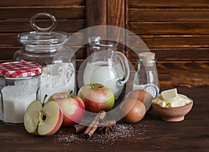 Ingredients and tools for baking - flour, eggs, butter, apples, cinnamon on a brown rustic wooden surface.