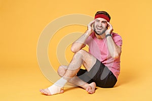 Ingured crying bearded fitness guy sportsman in headband t-shirt in home gym isolated on yellow background. Workout