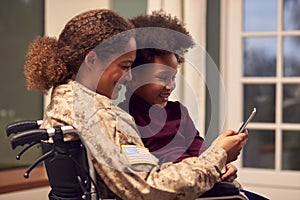 Injured Female American Soldier Wearing Uniform Sitting In Wheelchair Looking At Phone With Daughter