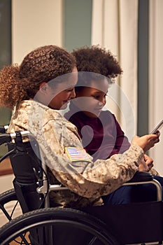 Injured Female American Soldier Wearing Uniform Sitting In Wheelchair Looking At Phone With Daughter