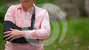 Injured mature woman arm sling suffering pain sitting outdoors, elbow fracture