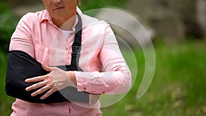 Injured mature woman arm sling suffering pain sitting outdoors, elbow fracture