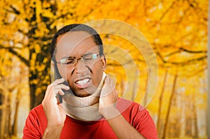 Injured young positive black hispanic male wearing neck brace, talking on phone and hand holding support in agony