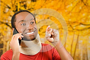 Injured young positive black hispanic male wearing neck brace and talking on phone smiling, yellow abstract background