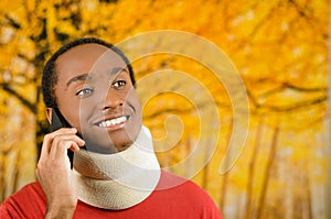 Injured young positive black hispanic male wearing neck brace and talking on phone smiling, yellow abstract background