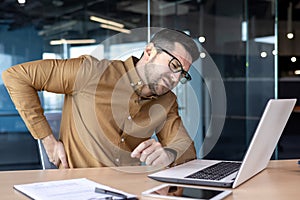 Injury at the workplace. A young man works in the office at a table behind a laptop and holds his hand behind his back