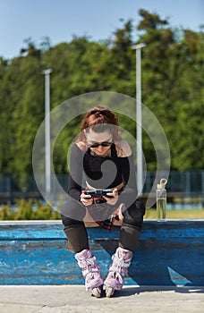 Inline skater female sitting on a ledge in a skatepark and playing online video game on a smartphone with a gamepad. Cool young