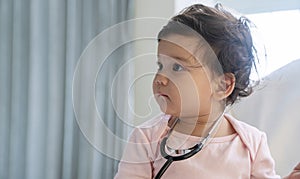 Innocence smile toddler girl sitting on the white bed with stethoscope while looking something at home. Mixed race little doctor