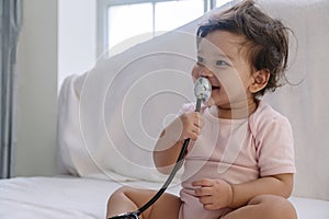 Innocence toddler girl sitting on the white bed while holding stethoscope in her hand at home. Mixed race little doctor baby