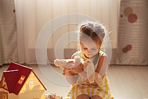 Innocently smiling girl with soft toy in hands sitting on floor with window on background, female child wearing yellow dress,