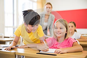 Inquisitive preteen boy peeking at workbook of cute smiling girl in class