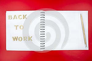 The inscription `back to work` made of wooden letters lying on an open notebook with white pages and a pencil, isolated on a red b