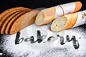 Inscription bakery on white wheat flour scattered Sliced french baguette and rye bread on dark background