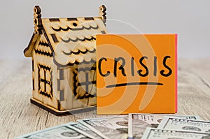 The inscription `crisis` on the sticker, dollars and a small wooden house. The concept of crisis, rising or falling real estate pr