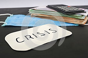 Inscription `crisis` on the sticker, medical mask, calculator and money stack on a black background. Crisis concept.